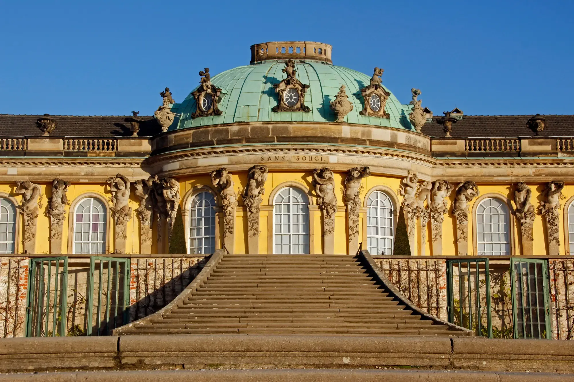 Yellow facade and green dome of Sanssouci Palace in Potsdam, with central stone stairs leading up.