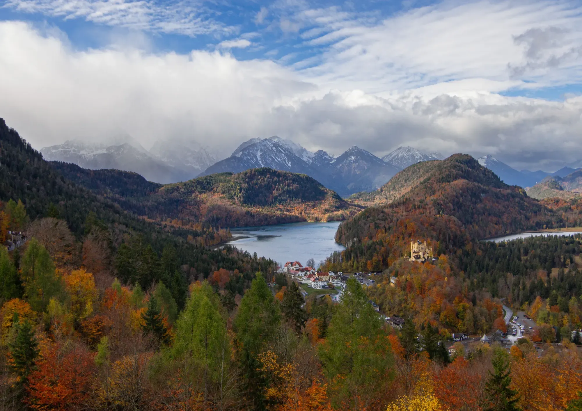 Hohenschwangau Castle and the Alpsee lake surrounded by autumn forests, with the Bavarian Alps in the background.