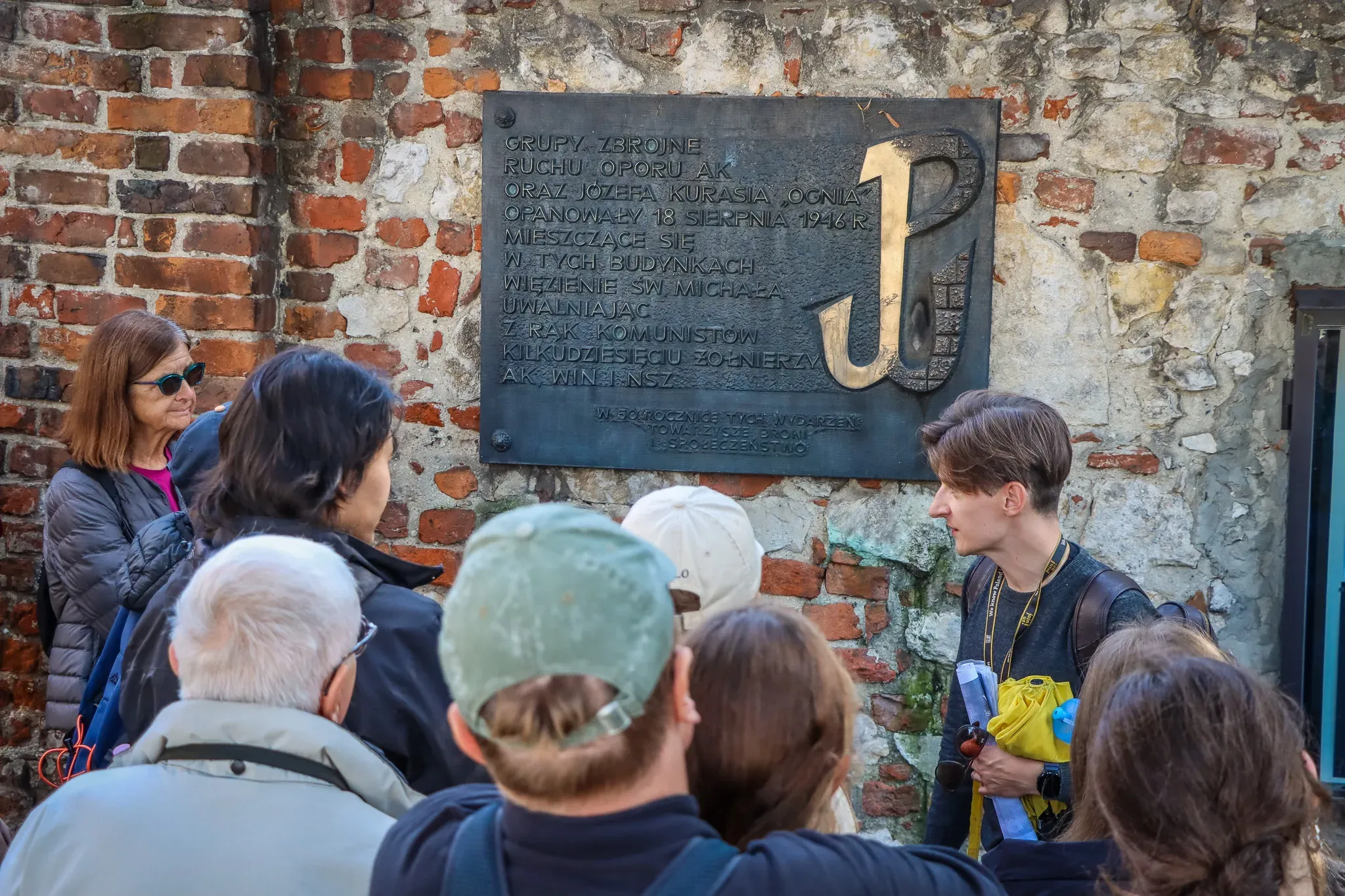 A tour guide explains a historical plaque with the Polish Fighting Symbol to a group of tourists against a brick wall.