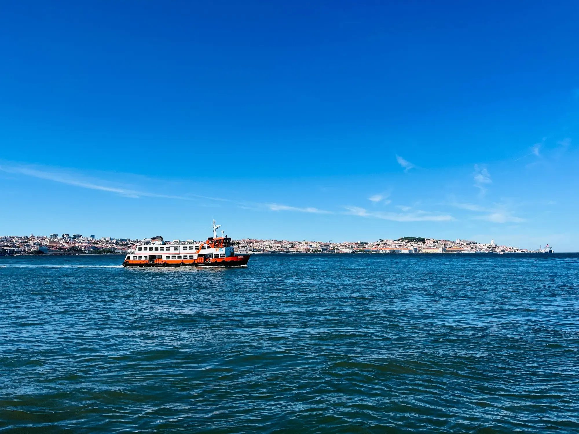 An orange and white ferry crosses the Tagus River in Lisbon, with the city skyline visible under a clear blue sky.