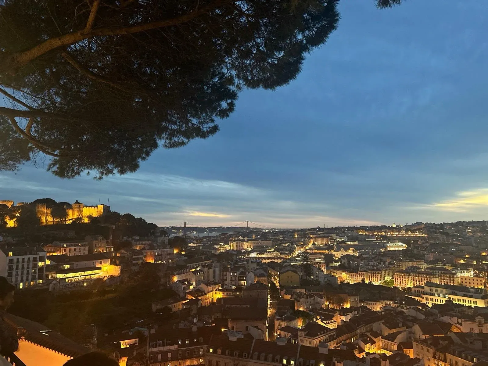 Lisbon's cityscape at dusk, with the illuminated São Jorge Castle on a hill and the 25 de Abril Bridge in the distance.