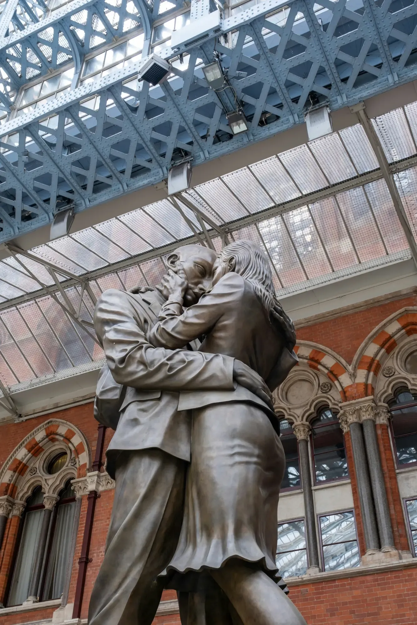 A giant bronze statue of a man and woman embracing under the arched glass roof of St Pancras station.