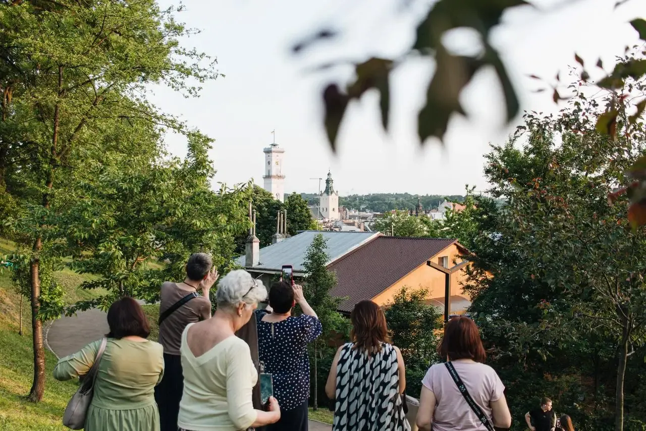 Group of people on a hill overlooking Lviv cityscape with City Hall tower and a church dome visible through trees.