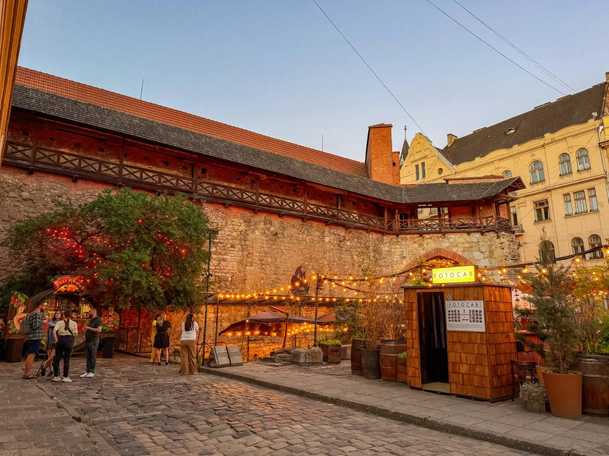 A cobblestone courtyard in Lviv at dusk, next to an old stone wall with a wooden gallery and an outdoor bar area.