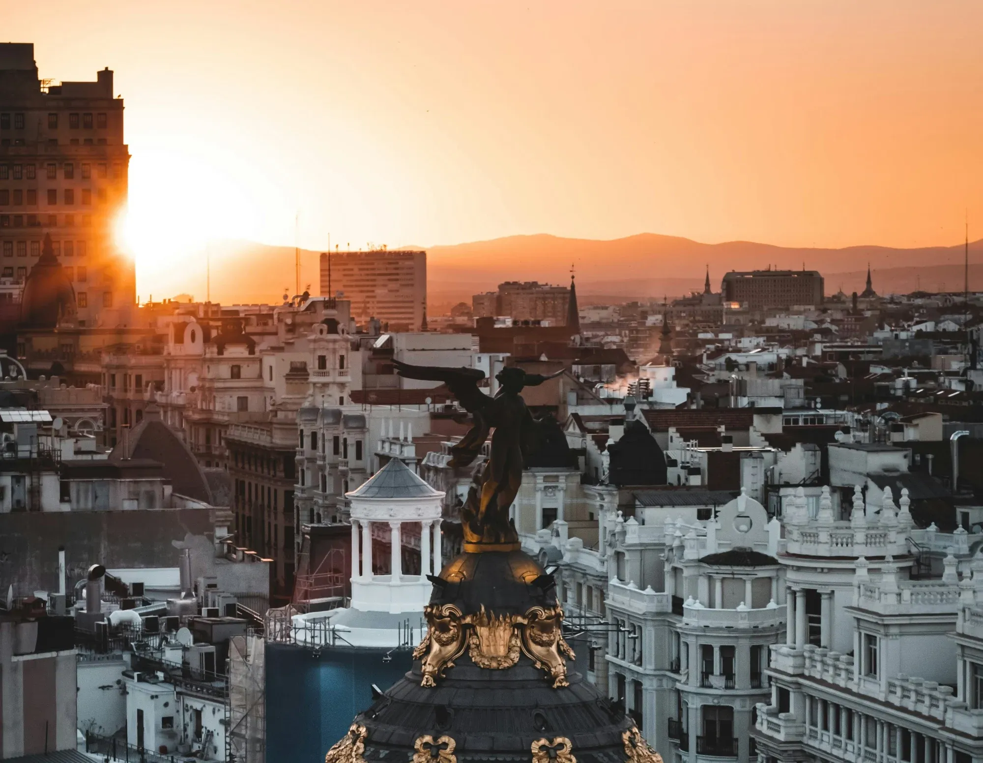 Rooftops of Madrid at sunset, with the dome of the Metropolis Building and a winged victory statue in the foreground.