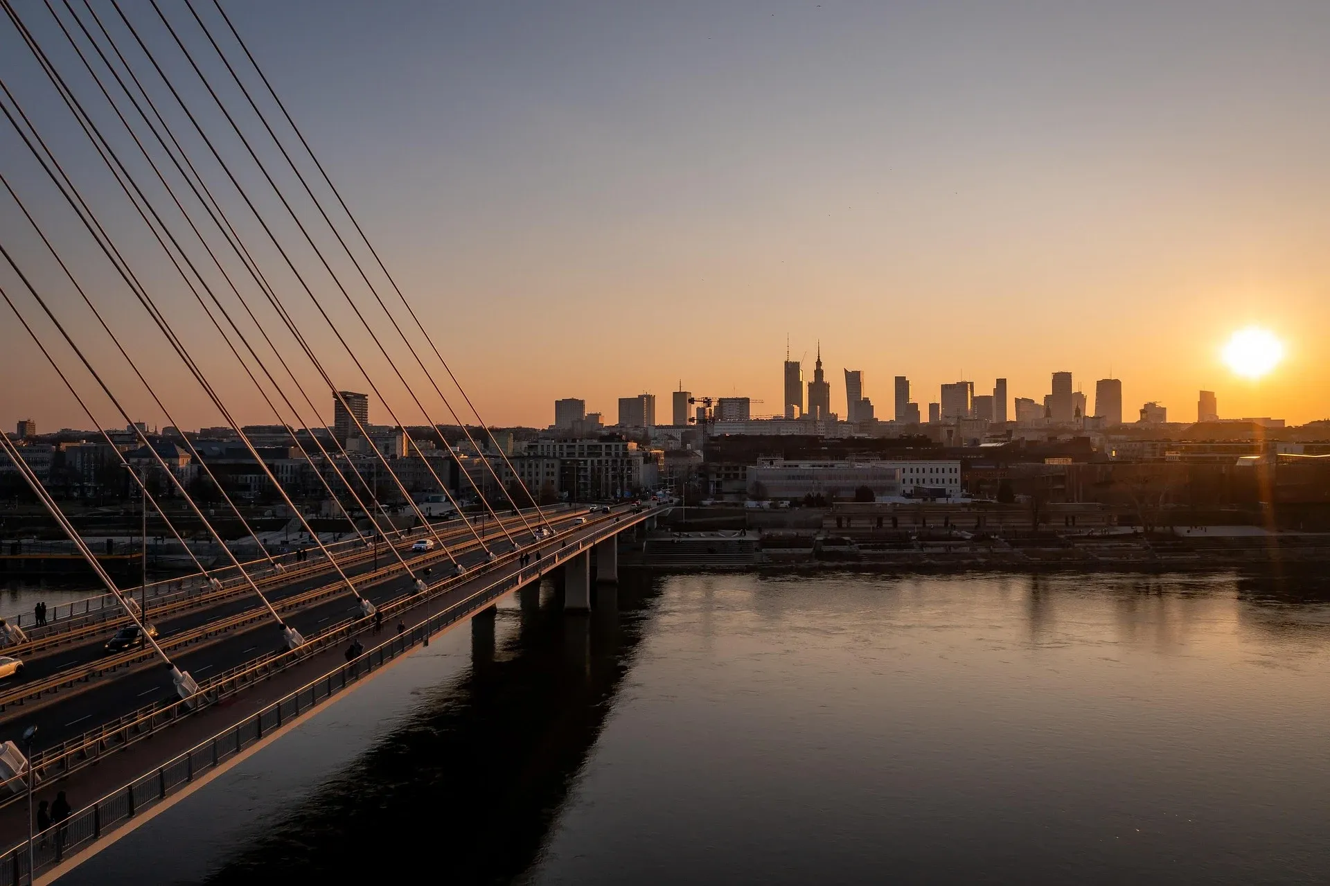 The Świętokrzyski Bridge over the Vistula River with the Warsaw city skyline against a golden sunset.