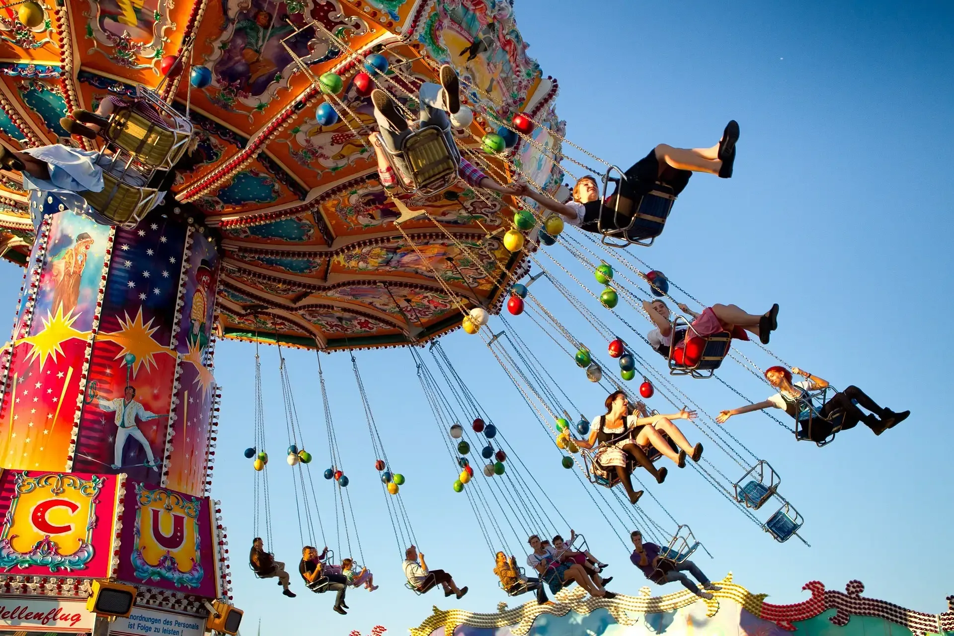 People riding a colorful chain swing carousel against a clear blue sky at a fairground, some wearing traditional Dirndls.