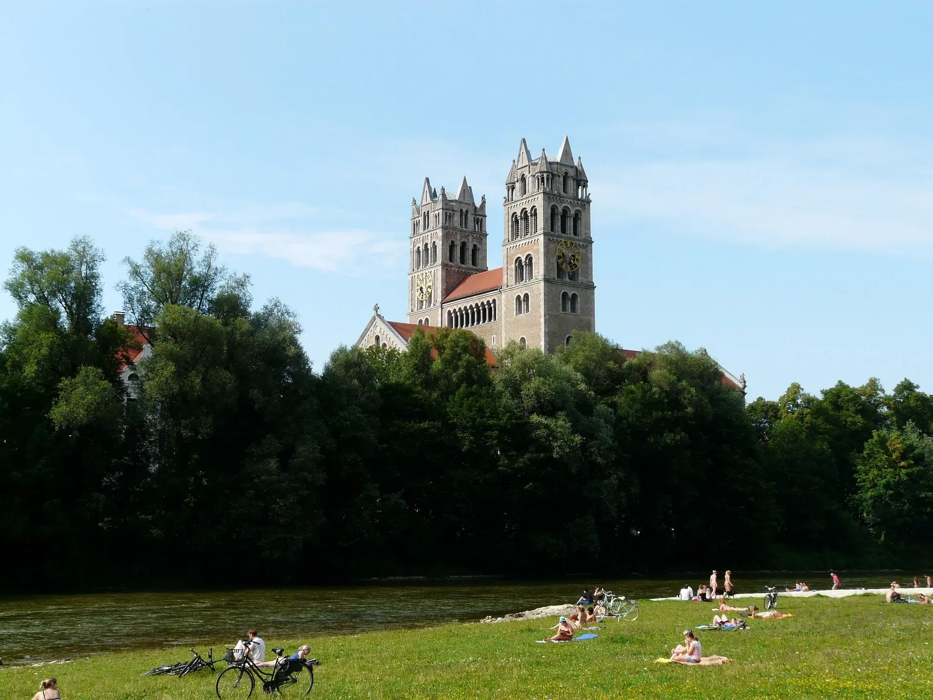 View of St. Maximilian church rising above trees, with people relaxing on the grassy banks of the Isar river in Munich.