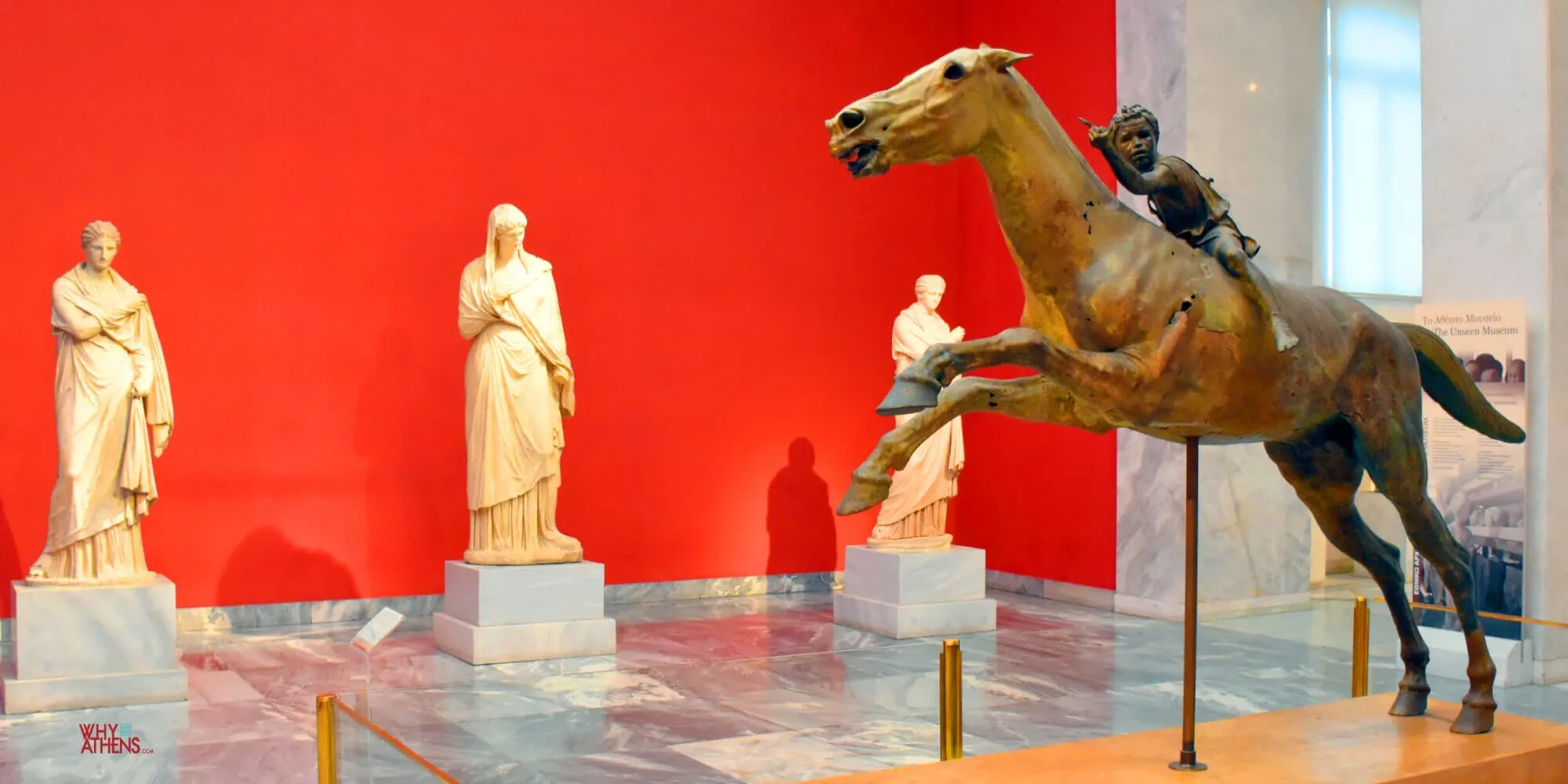 A bronze statue of a boy riding a galloping horse, displayed in a museum gallery with a bright red wall behind it.