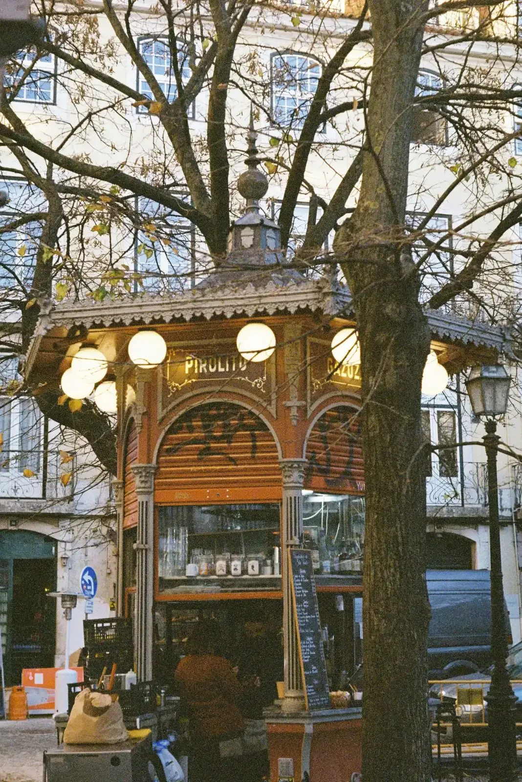 An ornate, old-fashioned street kiosk with round lights, seen through the bare branches of a tree in winter.