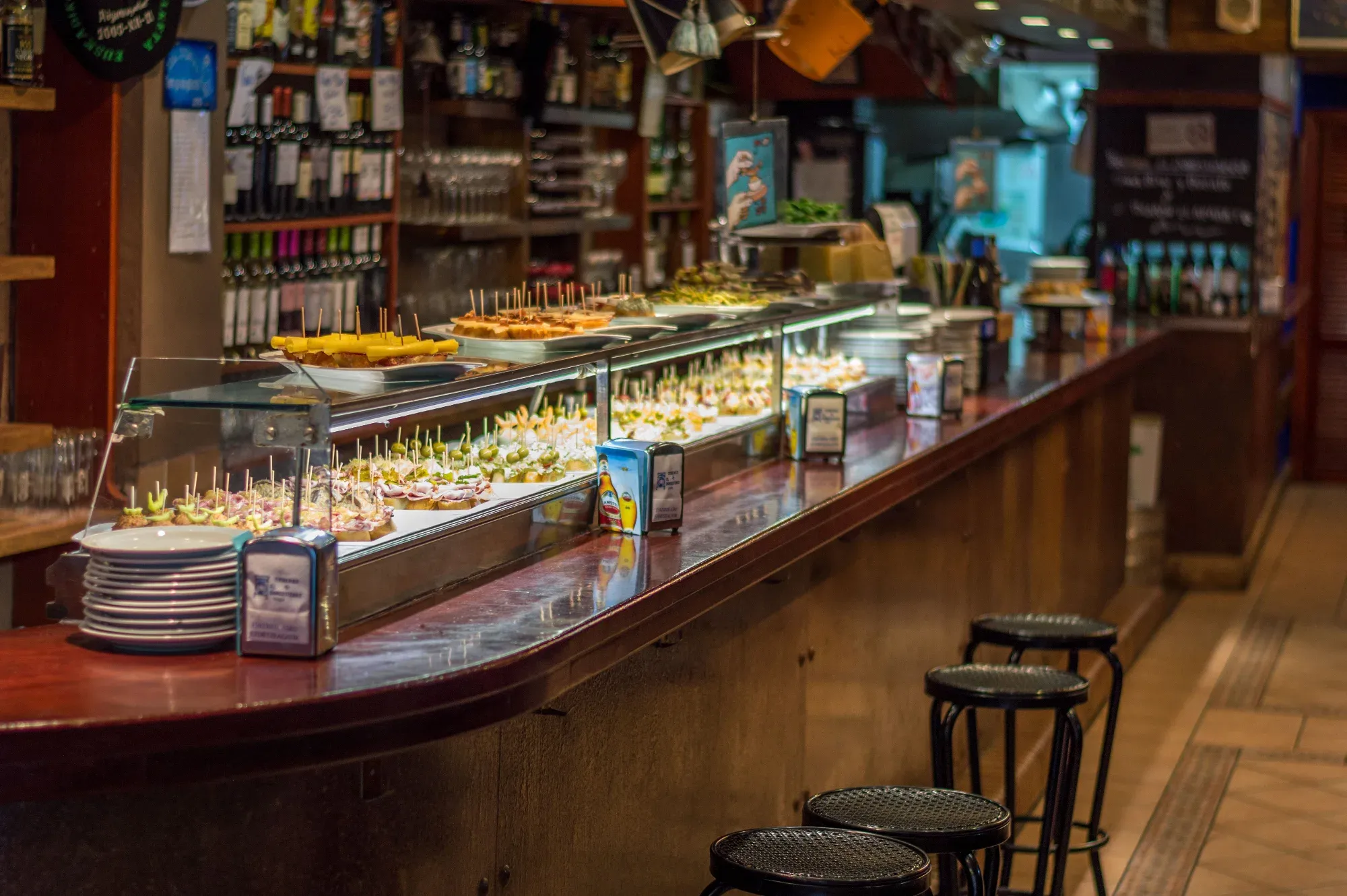 Assorted pintxos and tapas displayed along a long wooden bar counter with stools in a tavern.