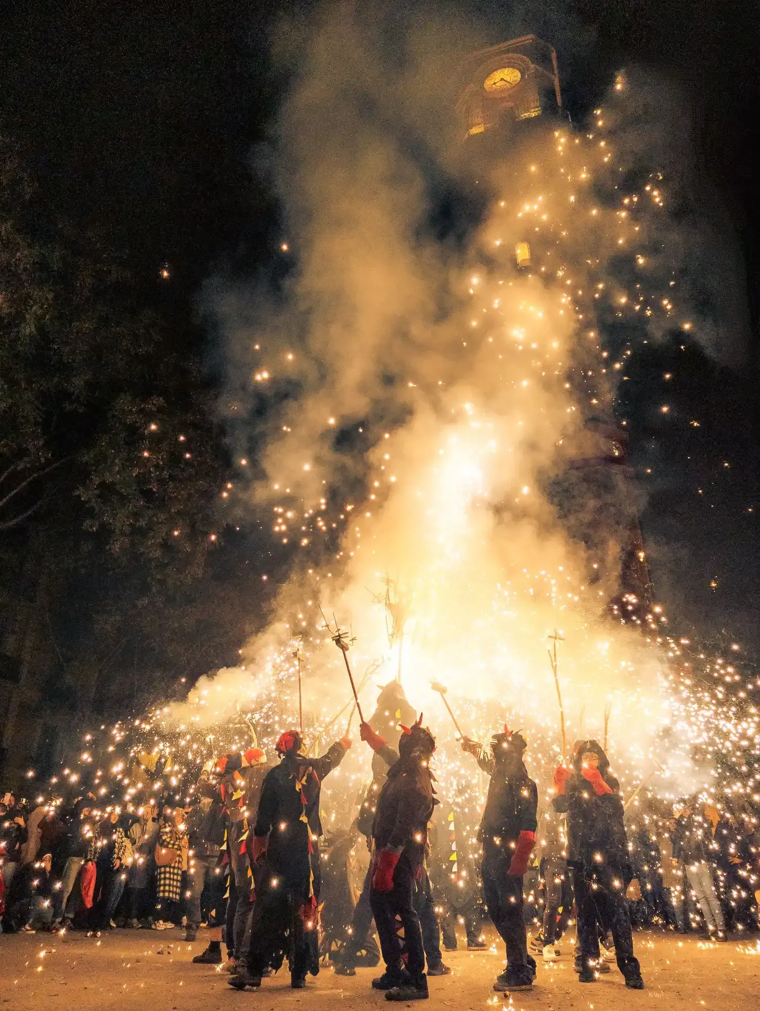 People in devil costumes hold staffs shooting sparks during a night street festival, with a crowd watching.