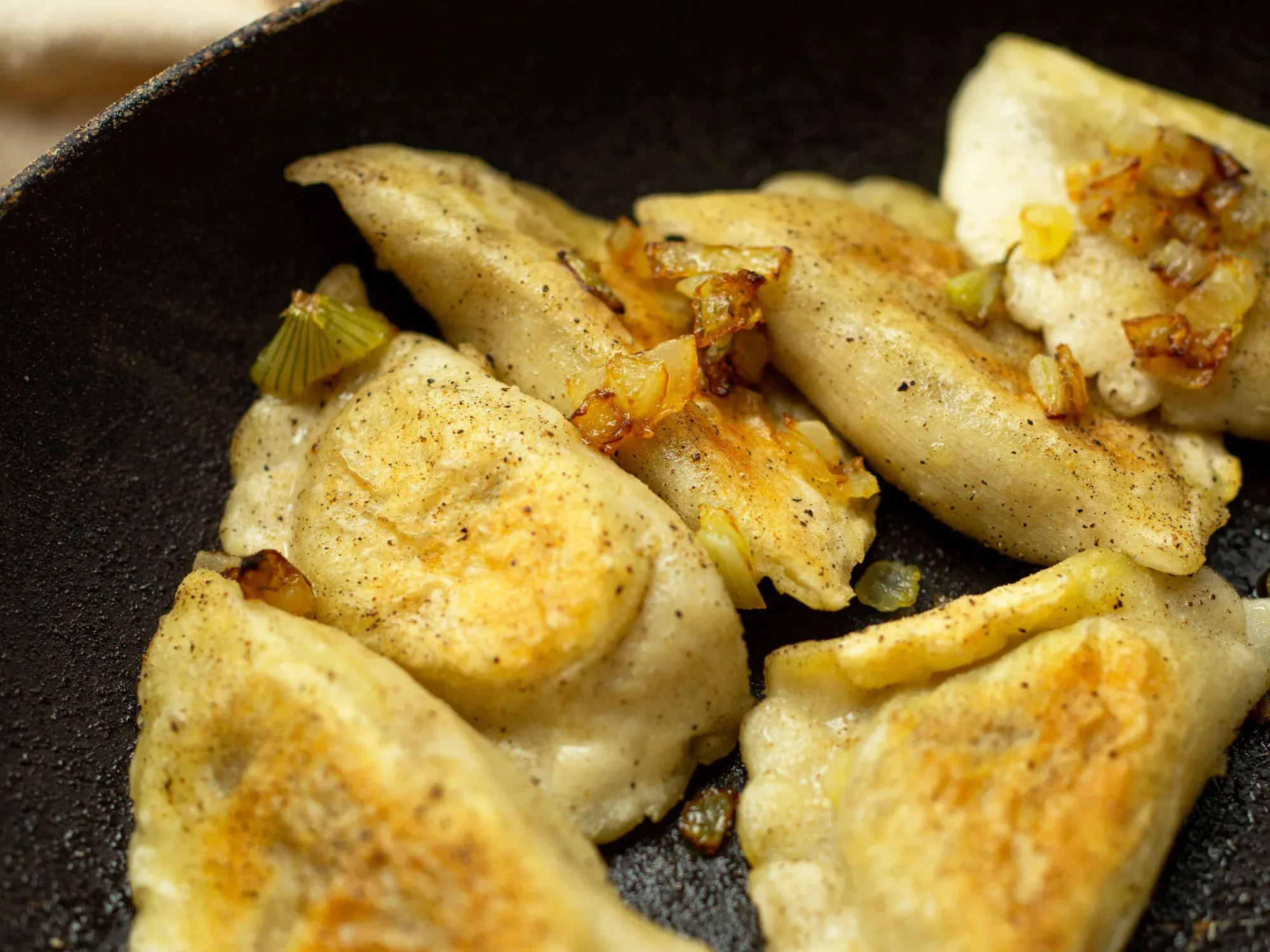 Close-up of pan-fried pierogi dumplings sprinkled with black pepper and topped with caramelized onions in a skillet.