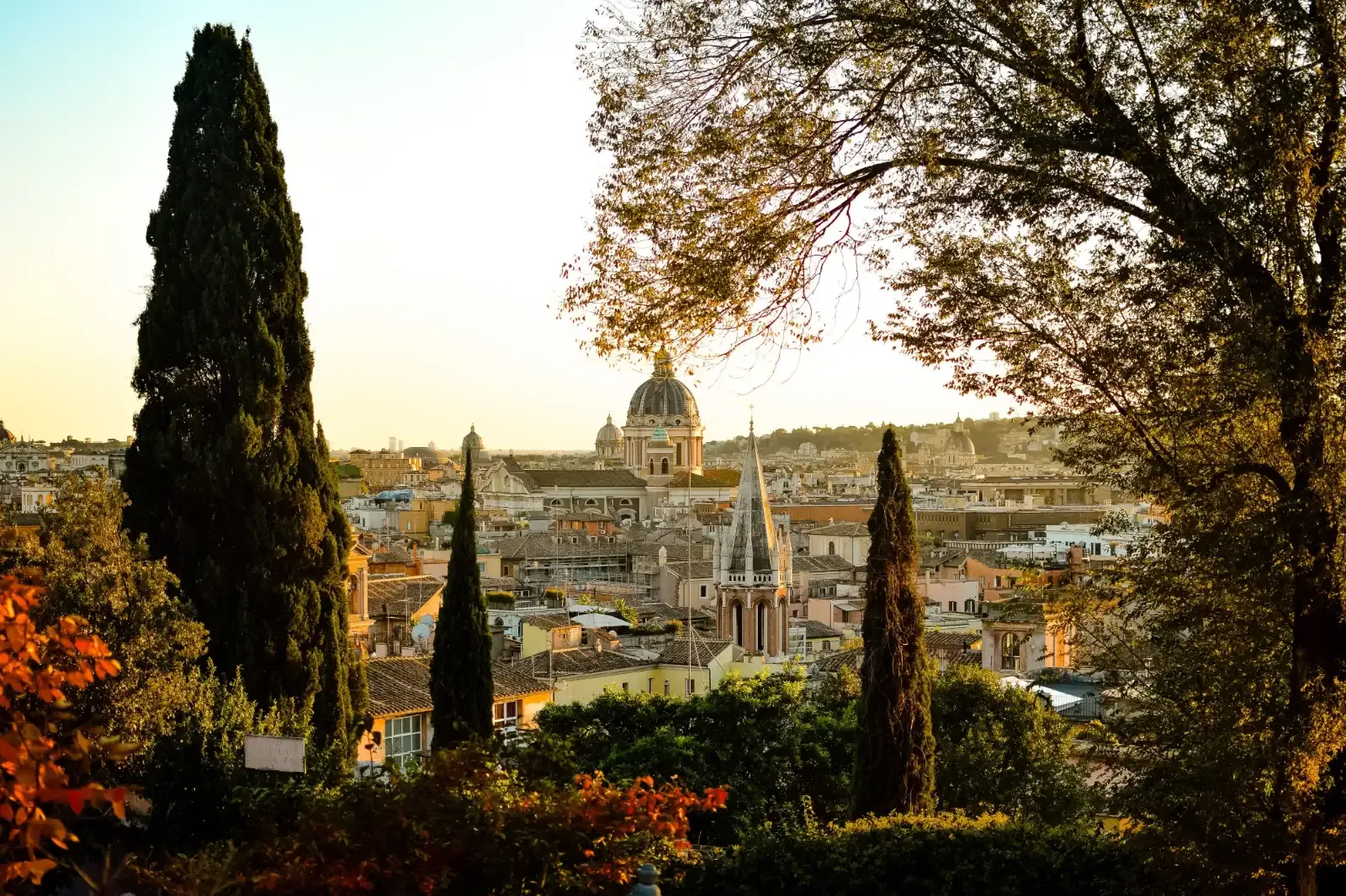 View over the rooftops and church domes of Rome at sunset, framed by cypress trees and foliage in the foreground.