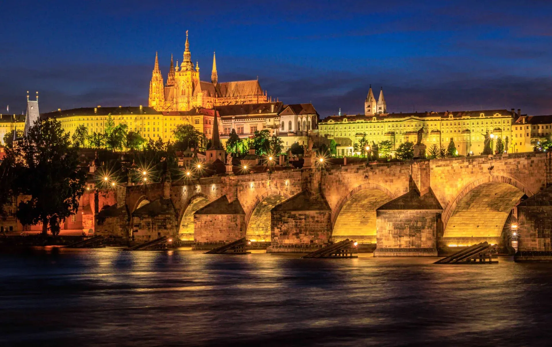 Illuminated Charles Bridge and Prague Castle at dusk, with the Vltava River reflecting lights below.