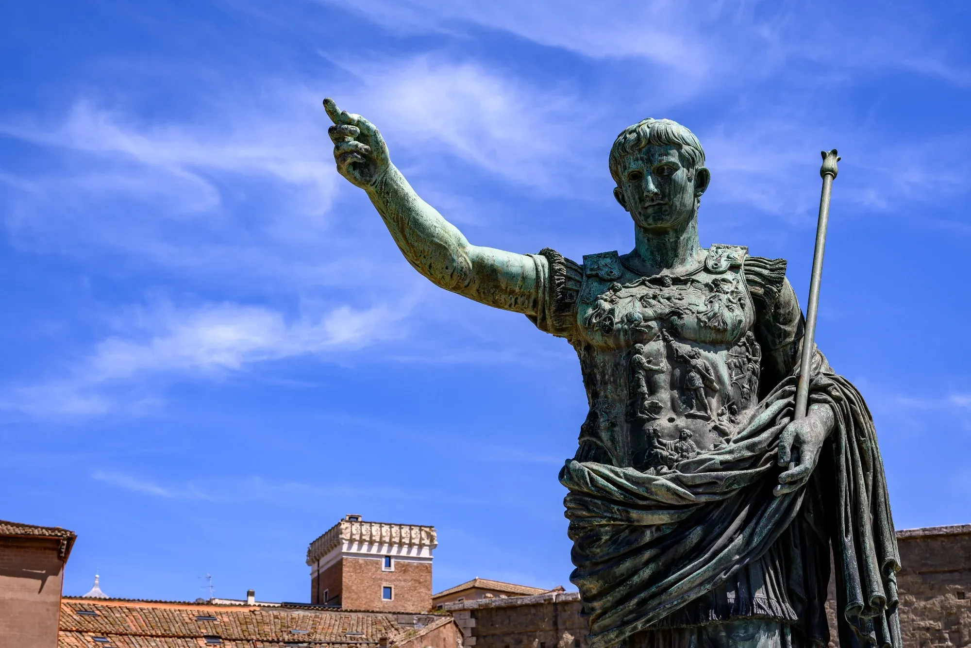 Bronze statue of Emperor Augustus in military dress against a blue sky, with Roman rooftops in the background.