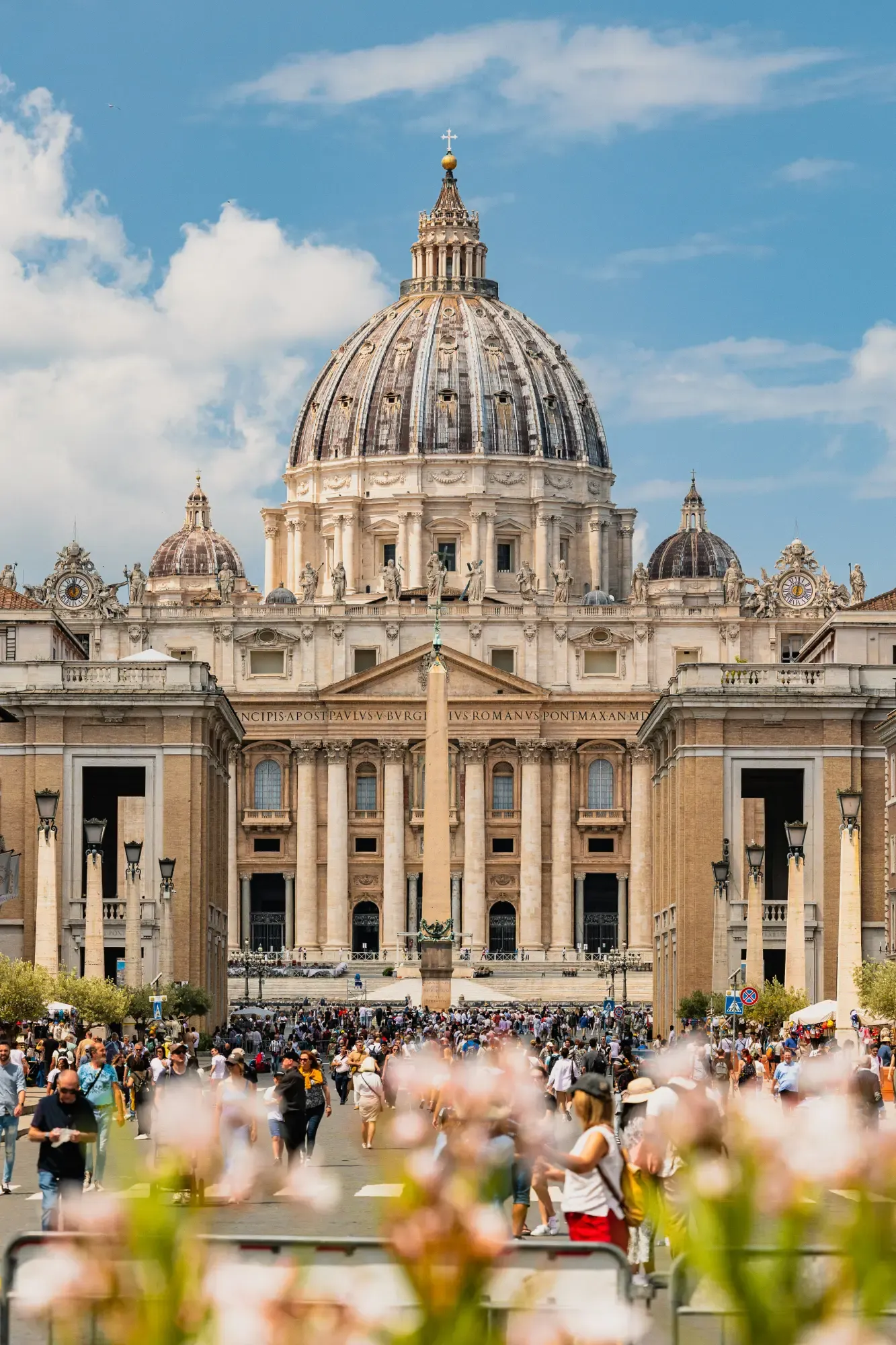 St. Peter's Basilica in Vatican City with a crowd of tourists in the square and blurred flowers in the foreground.