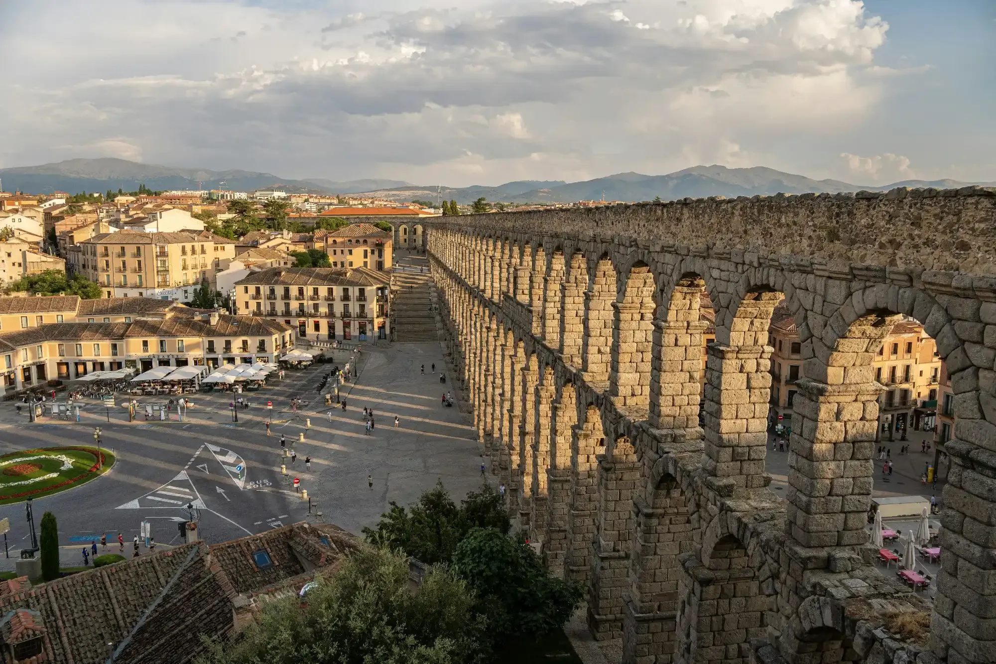 The Roman aqueduct of Segovia stretches across a city plaza, with buildings and mountains in the background.