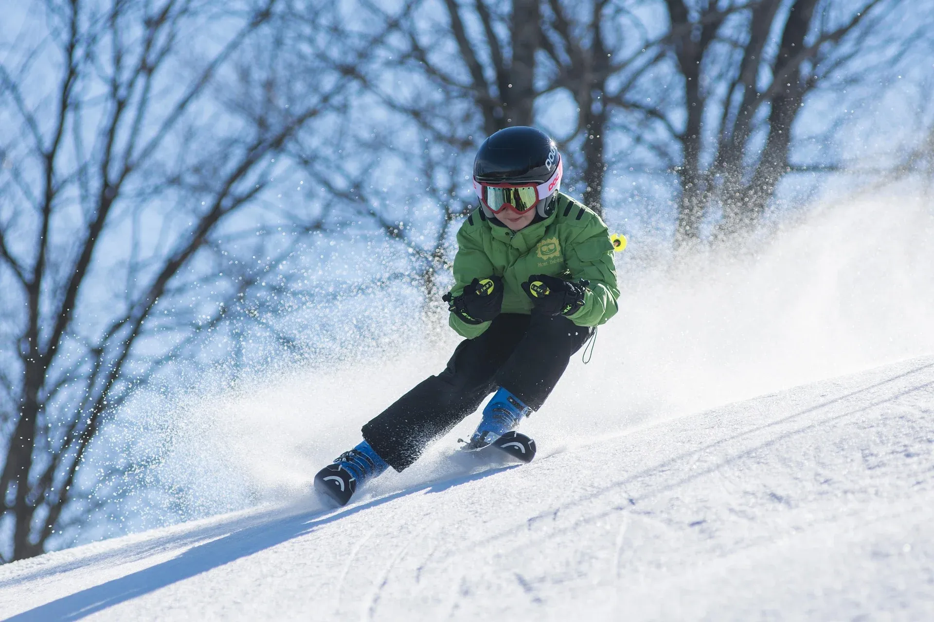 A child in a green jacket and helmet skiing down a sunny snowy slope, kicking up snow spray.