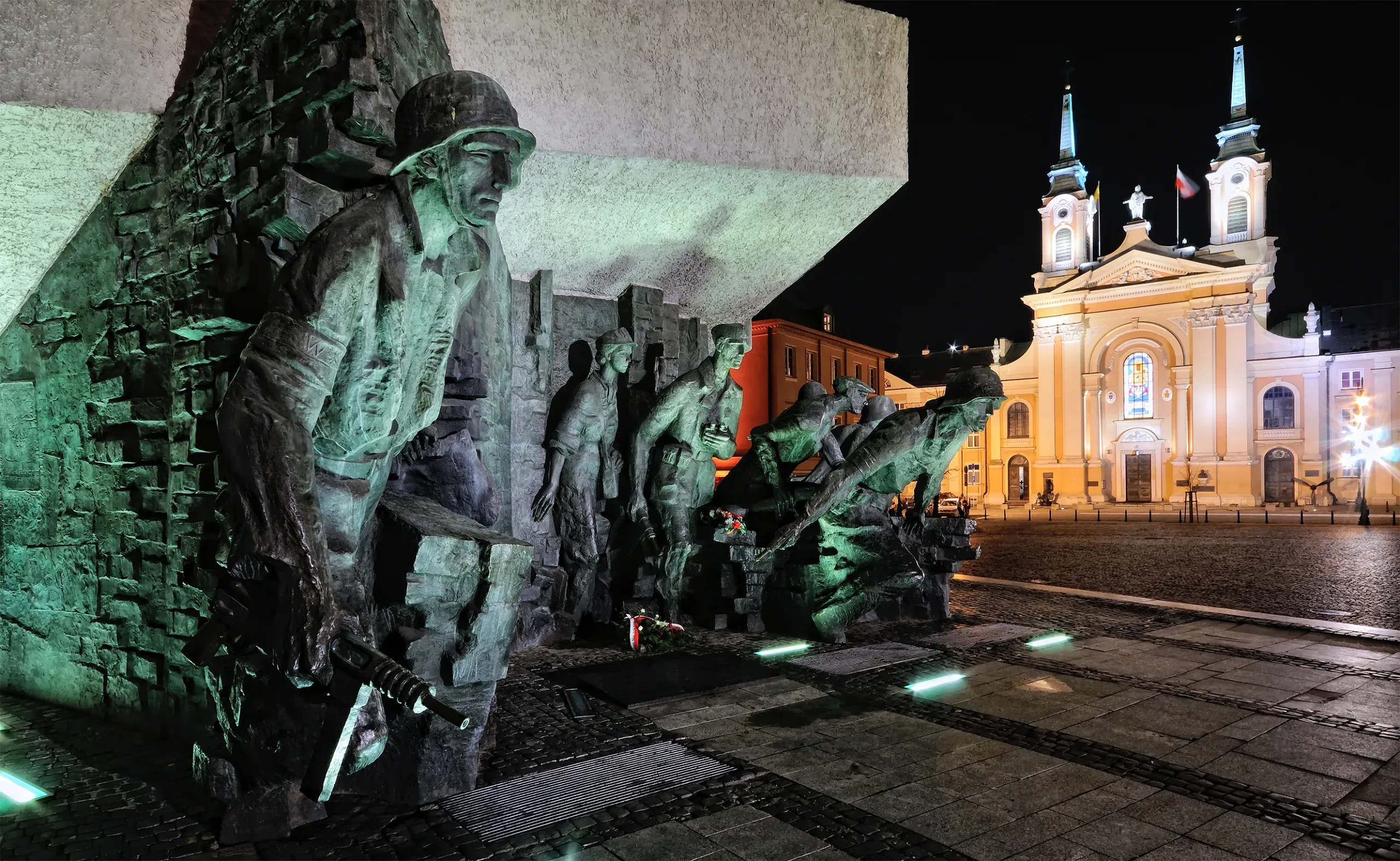 Bronze monument depicting figures of Warsaw Uprising soldiers under green light at night, with a brightly lit church behind.