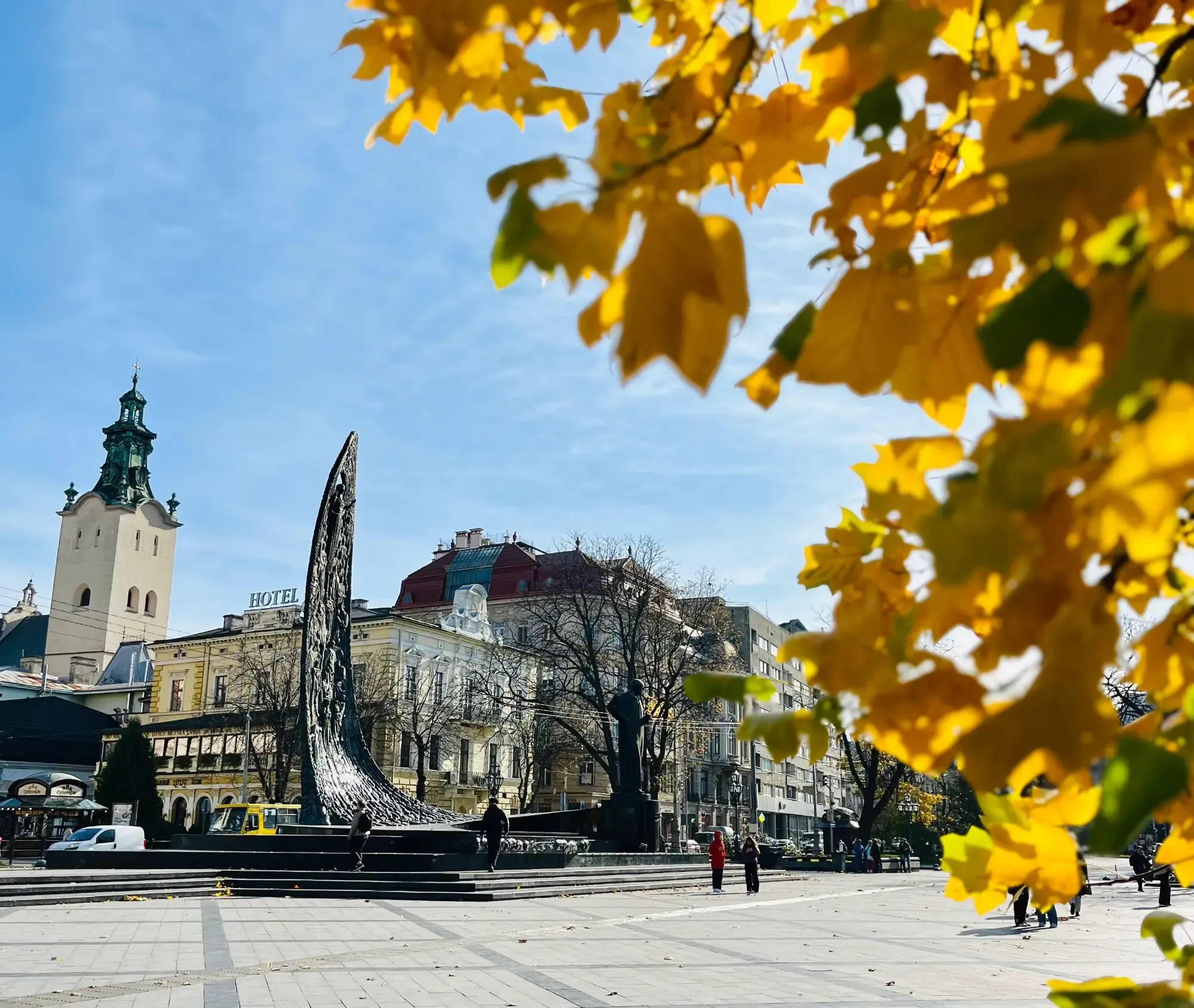 A sunny square in Lviv with the Latin Cathedral tower and a monument, seen through bright yellow autumn leaves.