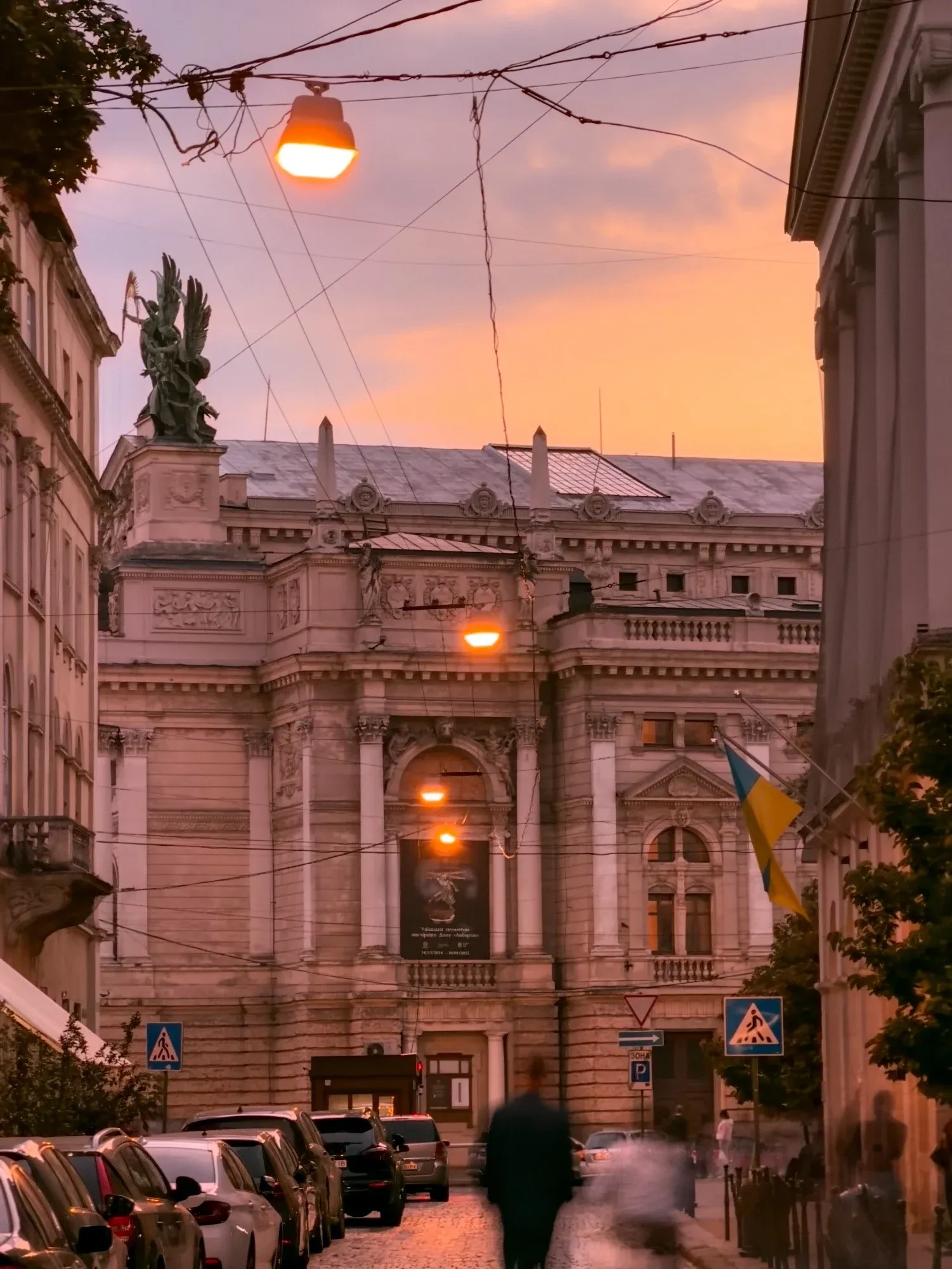 A street view of the Lviv Opera House at sunset, with warm streetlights and a Ukrainian flag.