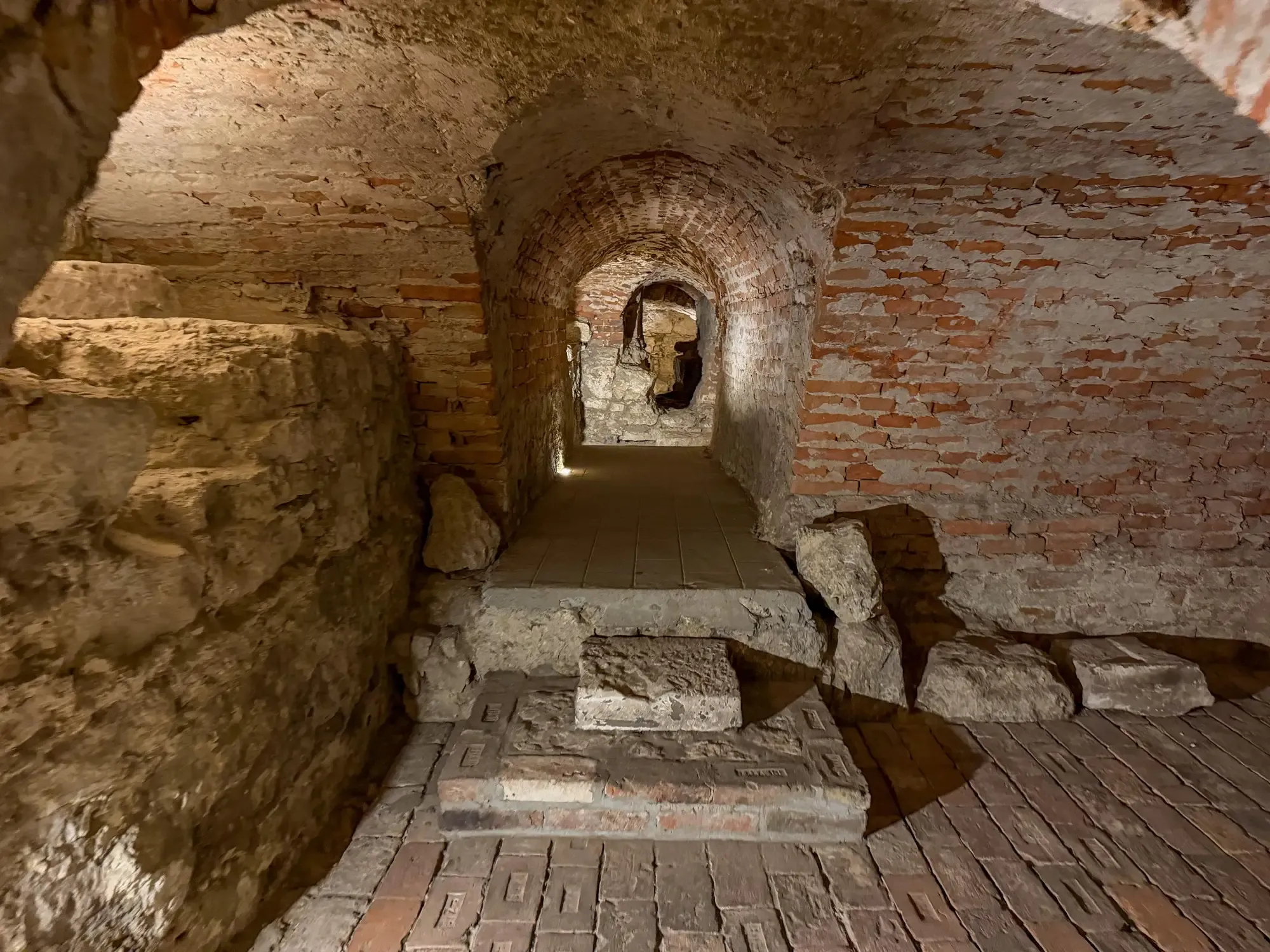 Dimly lit brick and stone vaulted underground corridor with low arched ceilings and stone steps.