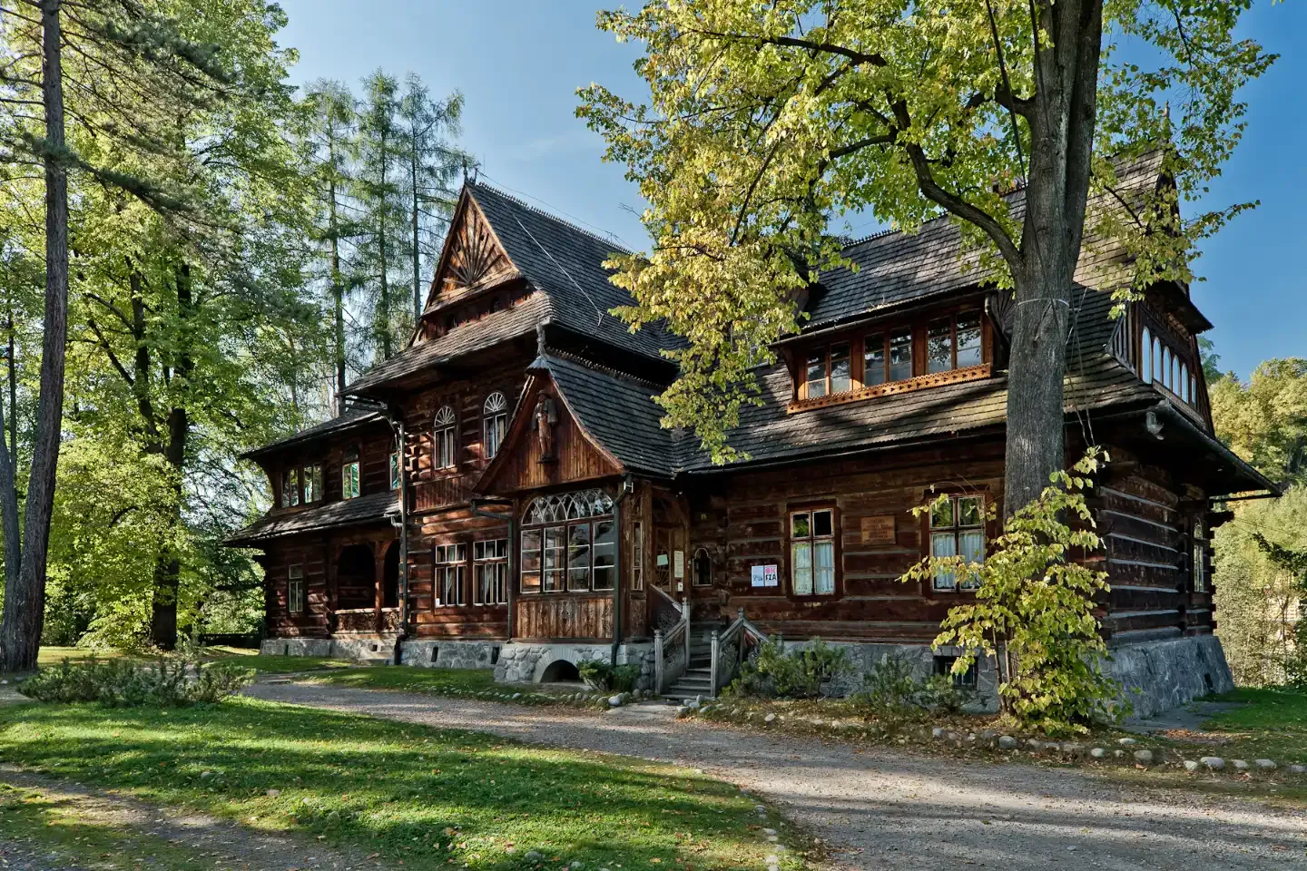 Two-story wooden villa with an ornate gabled roof and carved porch surrounded by trees on a sunny day.