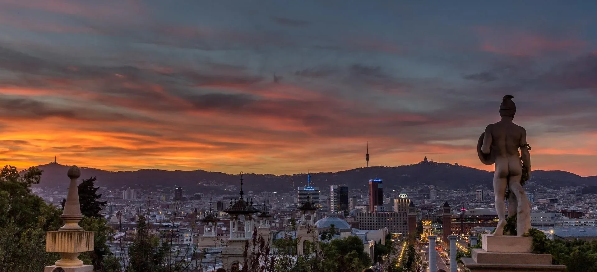 View of Barcelona city skyline at sunset with a statue in the foreground and Tibidabo mountain in the background.