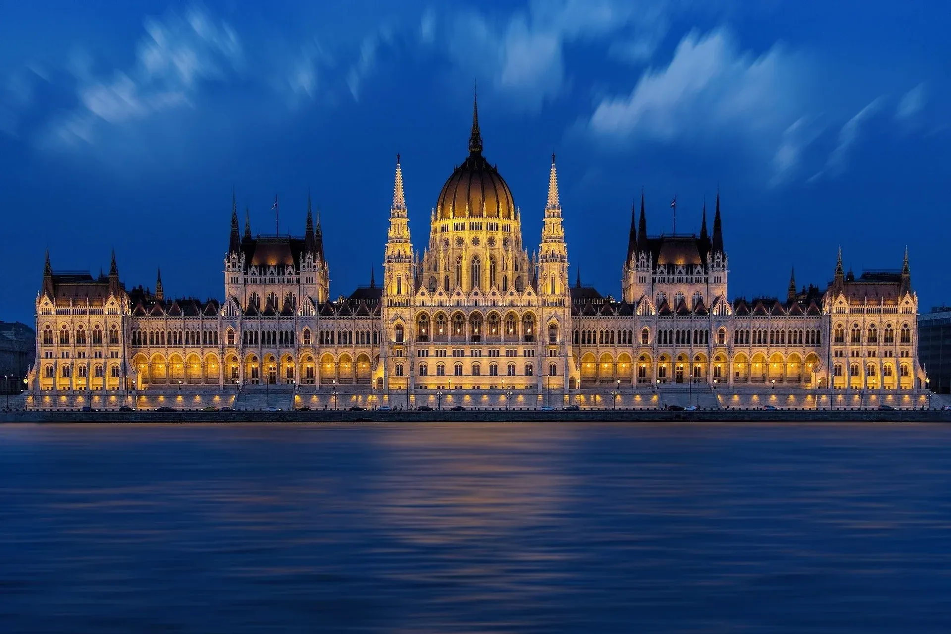 The illuminated Hungarian Parliament Building at night, viewed across the dark blue Danube River under a cloudy sky.