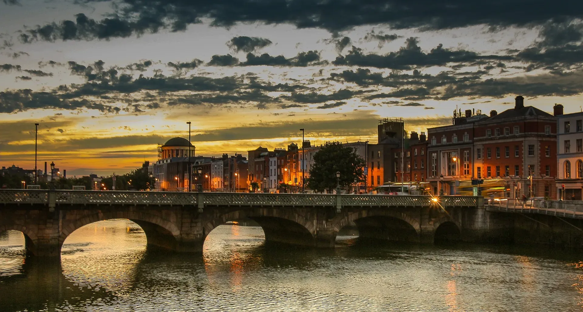 Stone arch bridge over a river in Dublin at sunset, with city buildings lit up on the far bank and a cloudy sky.