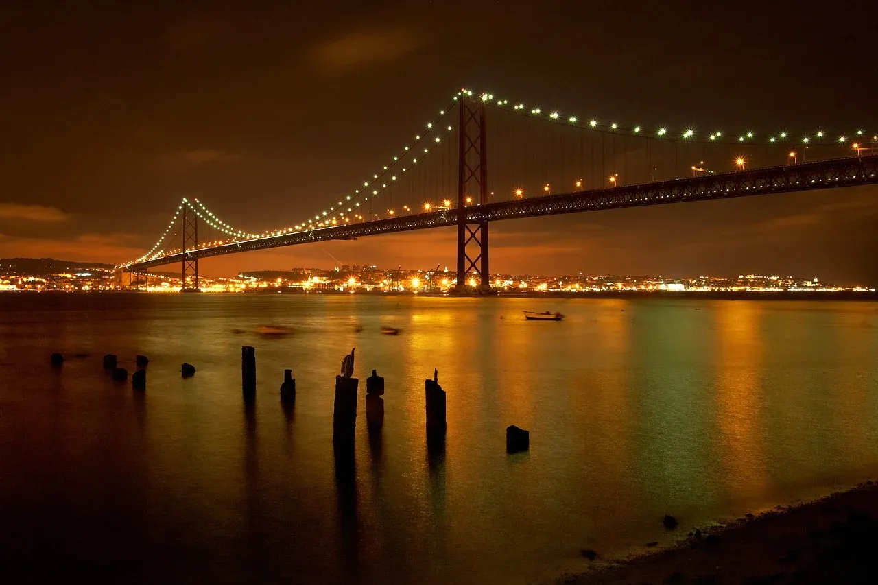 25 de Abril Bridge spanning a river at night, illuminated with city lights reflecting on the water. Wooden posts in foregroun