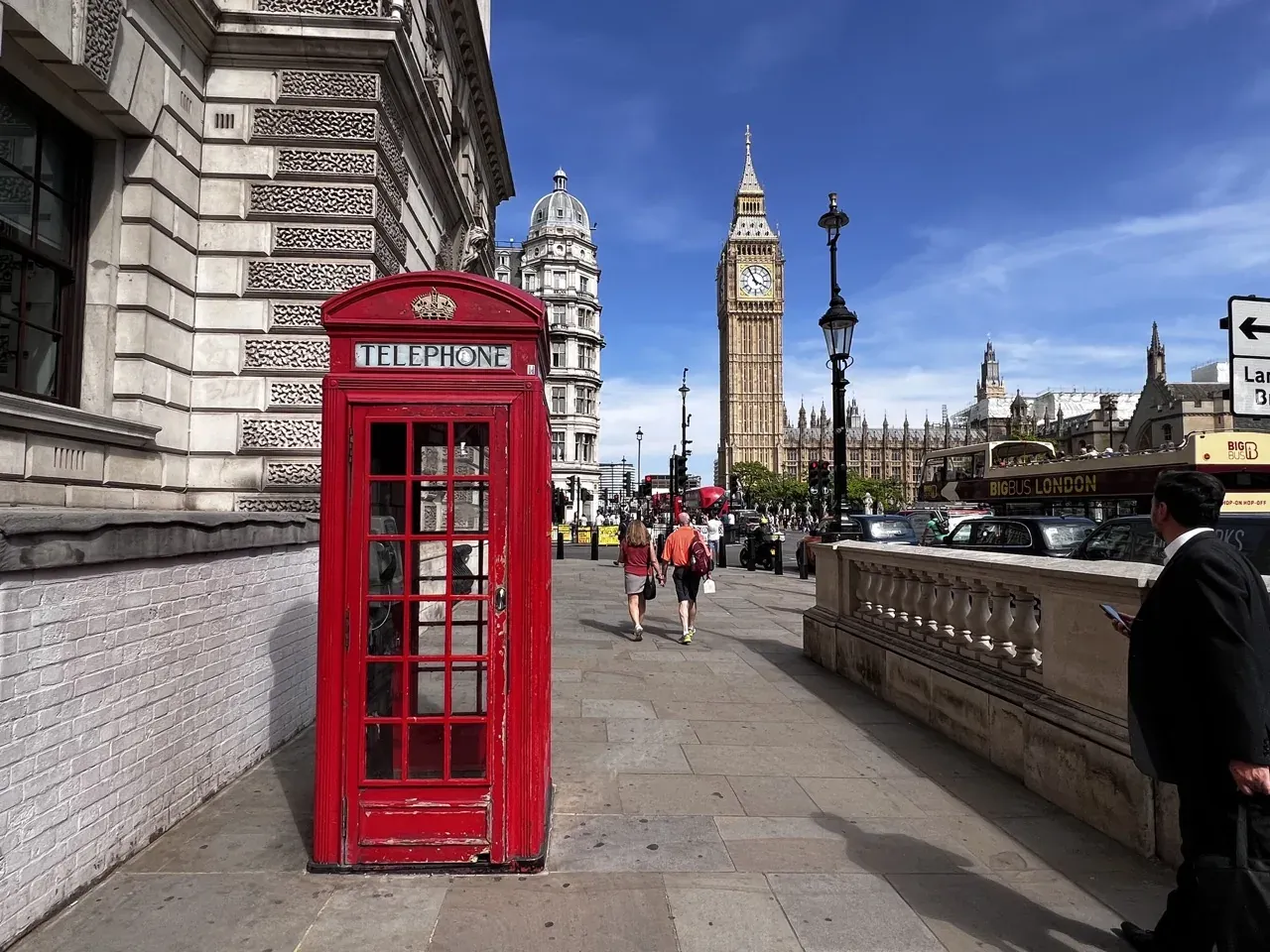 A classic red telephone booth on a London sidewalk with Big Ben and the Houses of Parliament in the background under a blue s