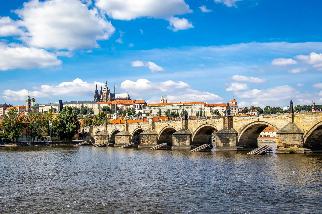 View of Charles Bridge with statues and arches spanning a river, Prague Castle on the distant left bank under a blue sky with