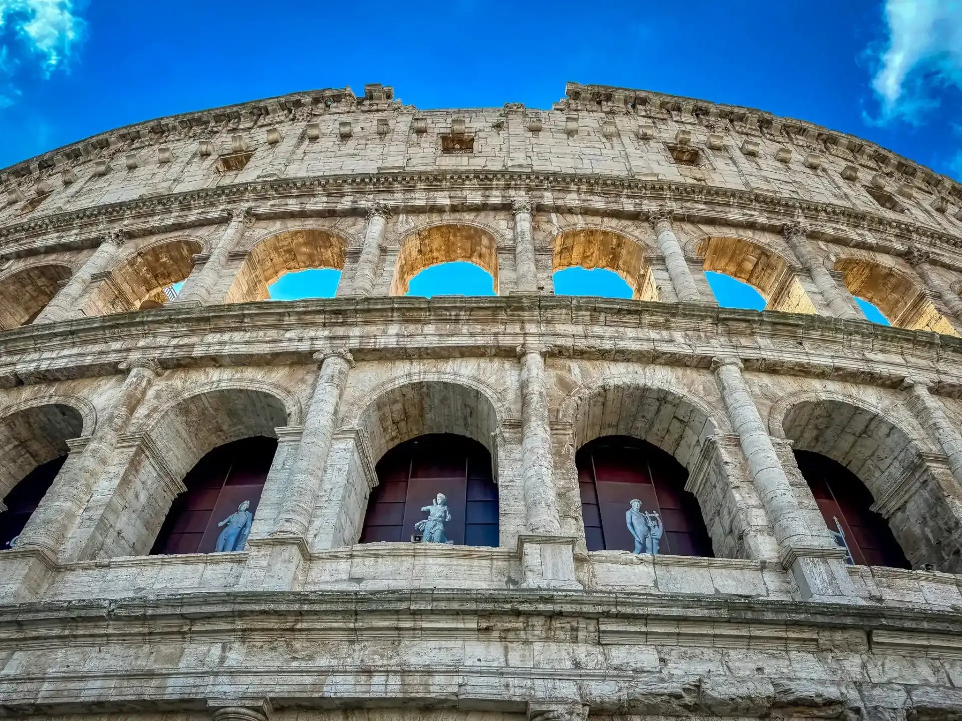 Exterior view of the Colosseum in Rome, showing multiple tiers of stone arches, columns, and statues in lower arches, against