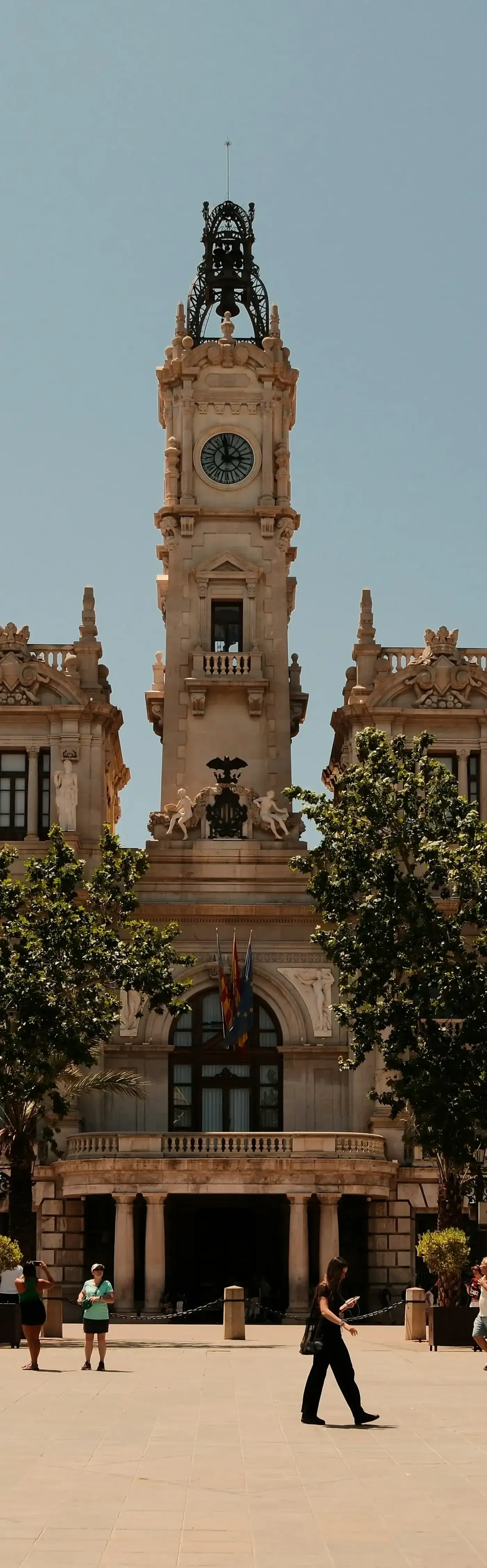 Ornate clock tower of Valencia City Hall with a large clock face, statues, flags, and green trees in foreground under a clear